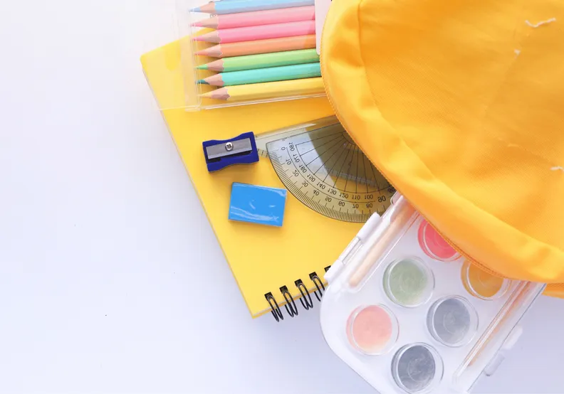 Primary school yellow backpack on otop of a white table with colourful school supplies comming out of it